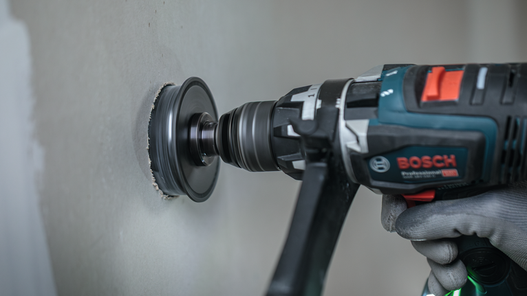 Person wearing safety equipment drills a circular hole into a drywall panel.