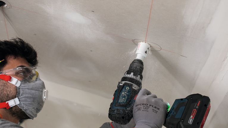 A person wearing safety equipment drills a circular hole in the ceiling.