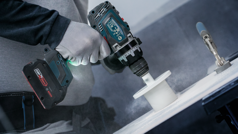 Person wearing safety equipment uses a cordless drill with a hole saw on a white surface.