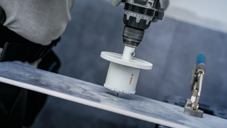 A person wearing safety equipment drills a hole into a ceramic tile.