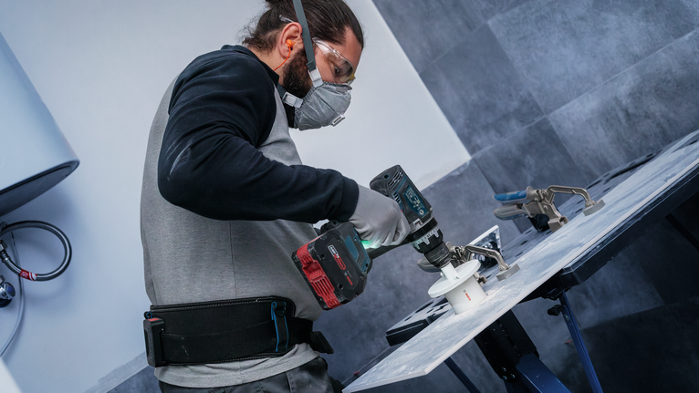Person wearing safety equipment drills a hole into a tile using a power tool.