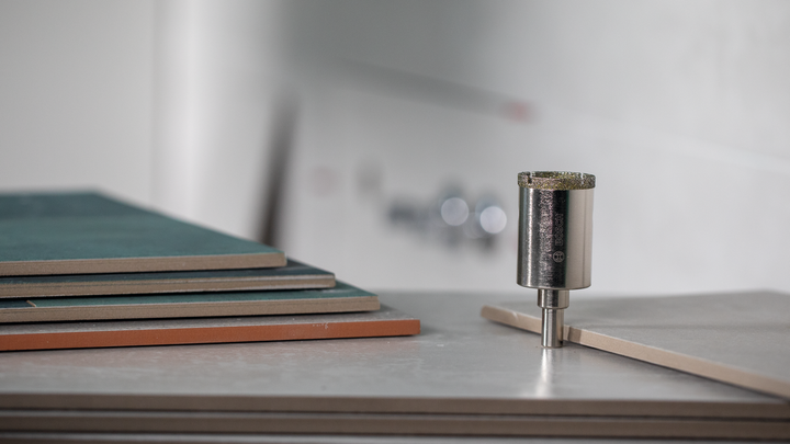 Diamond core drill bit positioned upright beside stacked tile sheets.