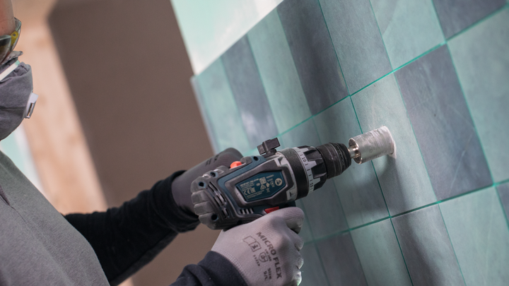 A person wearing safety equipment drills a hole into a tiled wall.