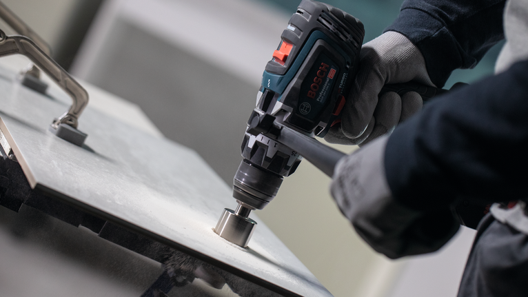 Person wearing safety equipment drills a hole into a tile surface.