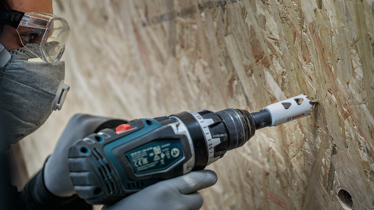 Person wearing safety equipment drills a hole into particle board.