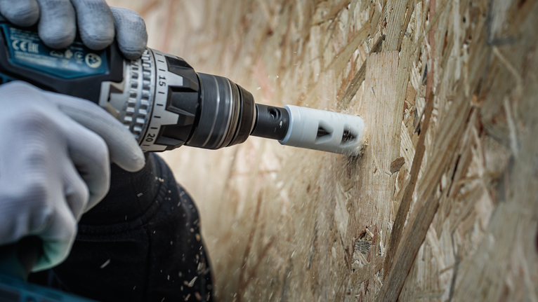 Person wearing safety equipment drills a circular hole in plywood.