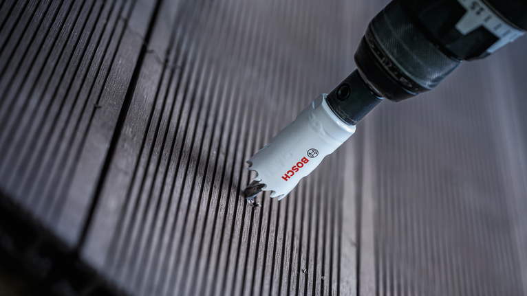 A hole saw drill bit creating a circular cut in a wooden deck plank.