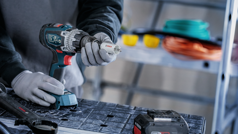 Person wearing safety equipment attaches a hole saw to a cordless drill on a workbench.