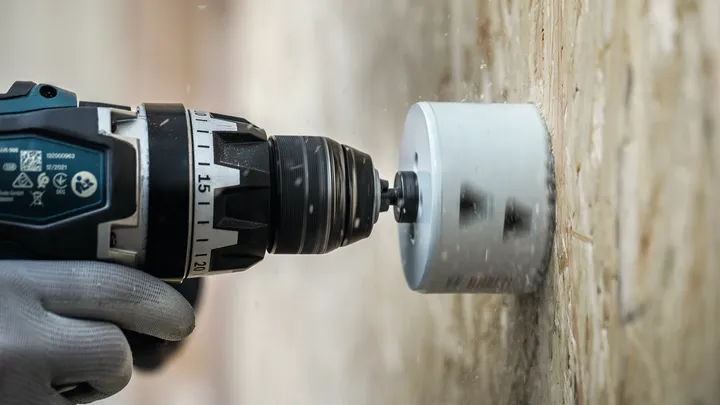 A person wearing safety equipment drills a large circular hole into a wooden surface.