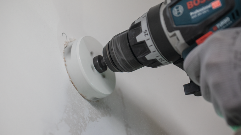 Person wearing safety equipment drills a circular hole into a wall.
