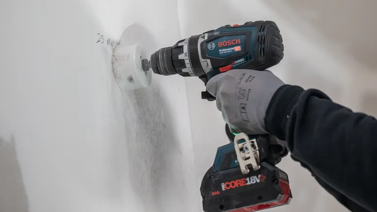 Person wearing safety equipment drills a circular hole into a white wall.