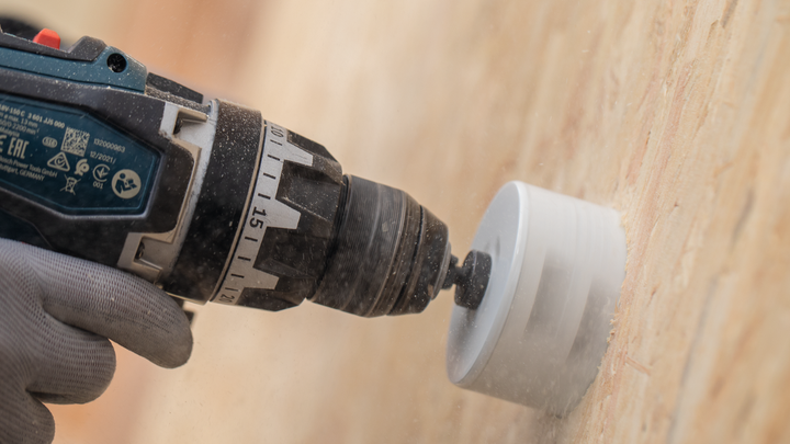 Person wearing safety equipment drills a circular hole in a wooden wall.