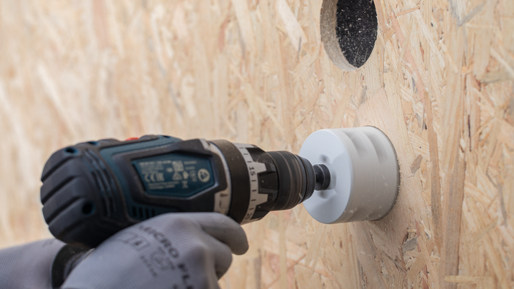 A person wearing safety equipment uses a drill with a hole saw to cut into a wood panel.