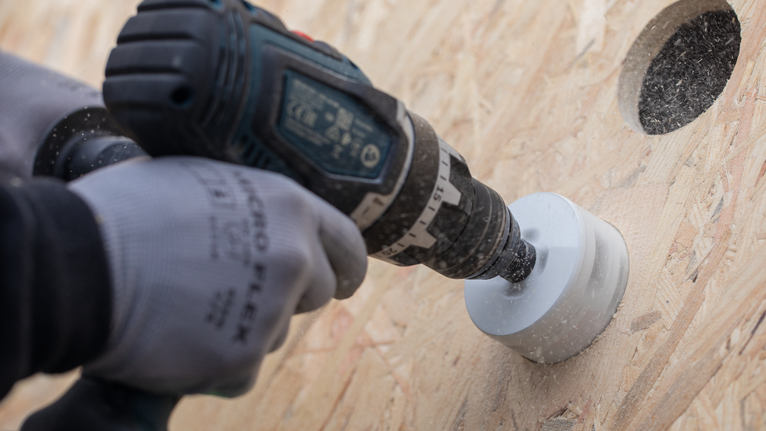 Person wearing safety equipment drills a circular hole in wood.