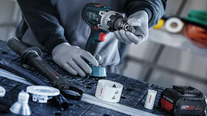 Person wearing safety equipment attaches a hole saw to a cordless drill on a workbench.