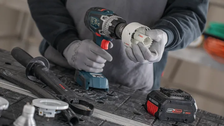 Person wearing safety equipment attaches a hole saw to a cordless drill on a workbench.