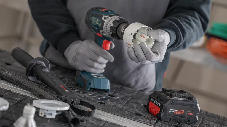 Person wearing safety equipment attaches a hole saw to a cordless drill on a workbench.