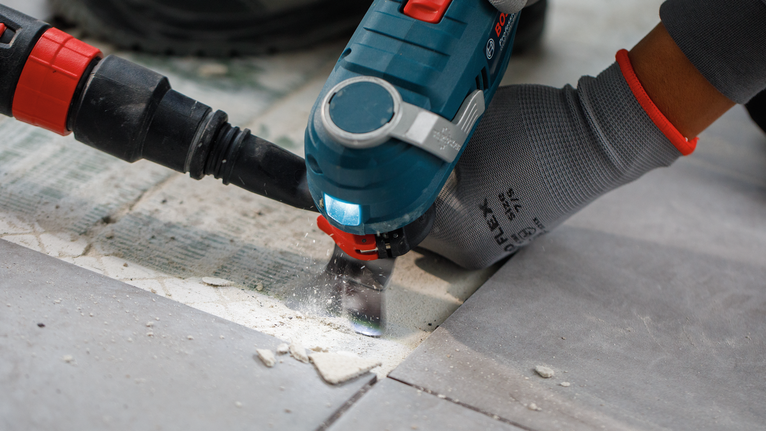 Person wearing safety equipment uses a power tool to cut through tile grout.