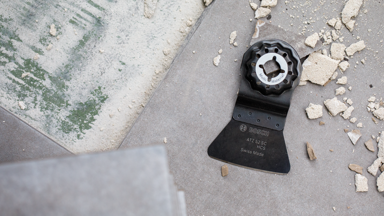 Oscillating multi-tool blade lies on a tiled floor surrounded by construction debris.