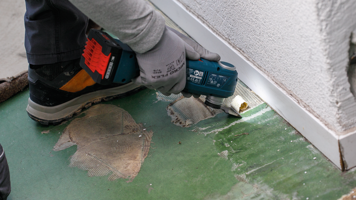 A person wearing safety equipment scrapes old adhesive from a floor near a wall.