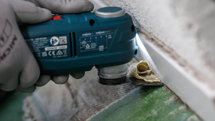 Person wearing safety equipment uses a power tool to cut pipe insulation near a wall.
