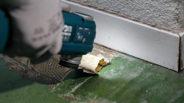 Person wearing safety equipment removes old flooring with a power tool near a baseboard.