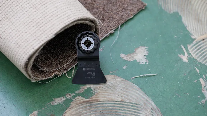 Oscillating tool blade attachment on a floor next to a rolled carpet.