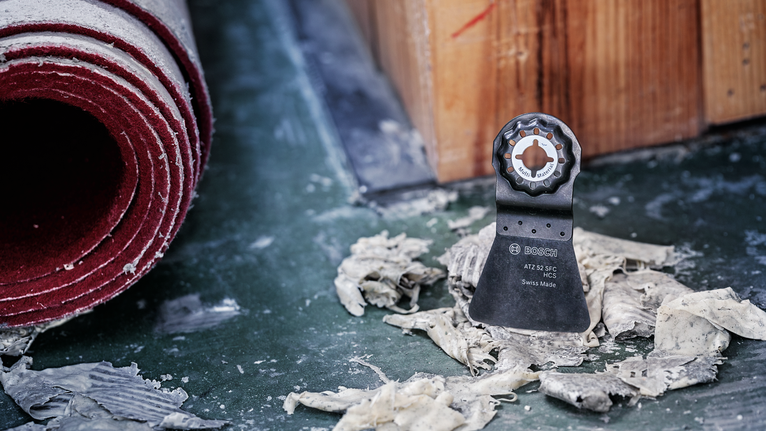 Oscillating tool blade stands upright on a dusty floor beside debris and a rolled carpet.