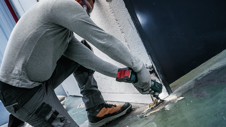 A person wearing safety equipment uses a power tool to remove flooring near a door.