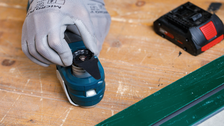 Person wearing safety equipment attaches a blade to a cordless multi-tool on a workbench.