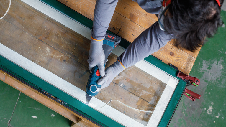 Person wearing safety equipment uses a power tool to cut a window frame.