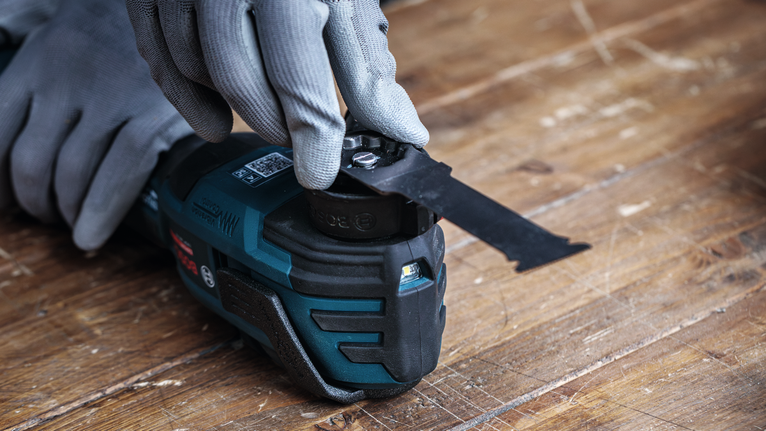 Person wearing safety equipment adjusts a multitool blade on a wooden surface.