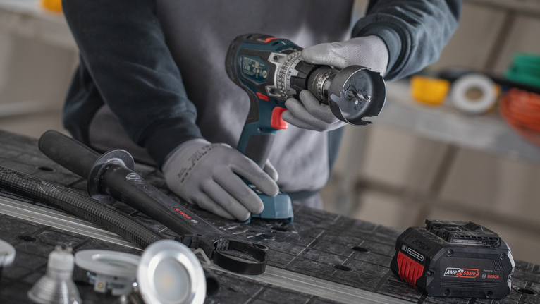 Person wearing safety equipment uses a cordless drill on a workbench.