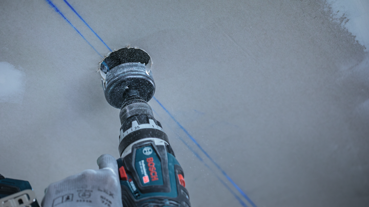 Person wearing safety equipment drills a circular hole in drywall.