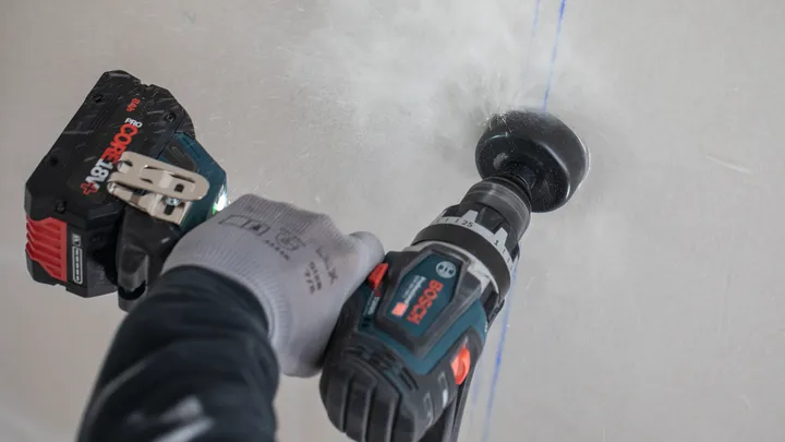 Person wearing safety equipment drills a hole into a white wall.