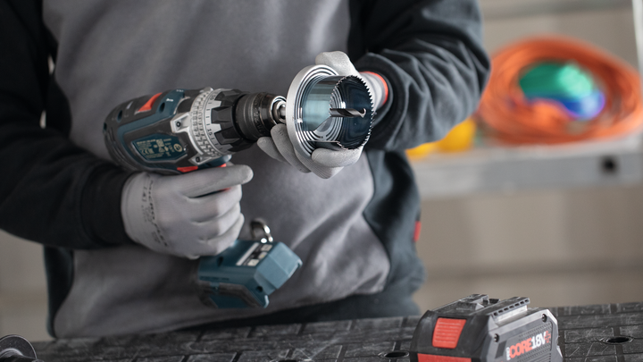 A person wearing safety equipment attaches a hole saw to a cordless drill.