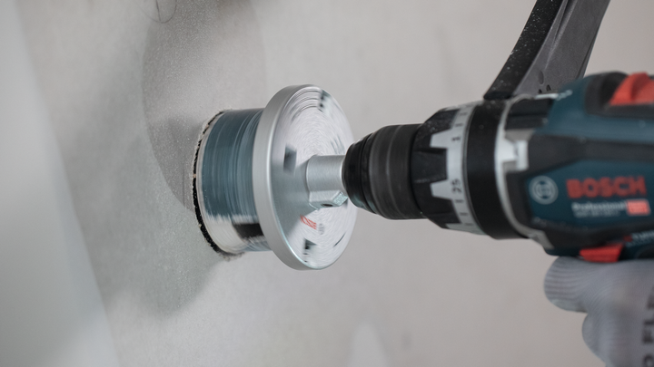 A person wearing safety equipment drills a large circular hole into a wall.