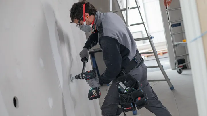 Person wearing safety equipment drills a hole into drywall in a construction area.