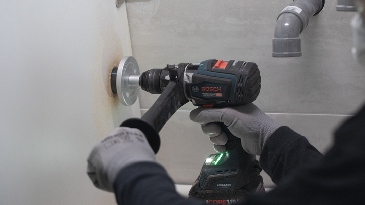 Person wearing safety equipment drills a hole in a tiled wall.