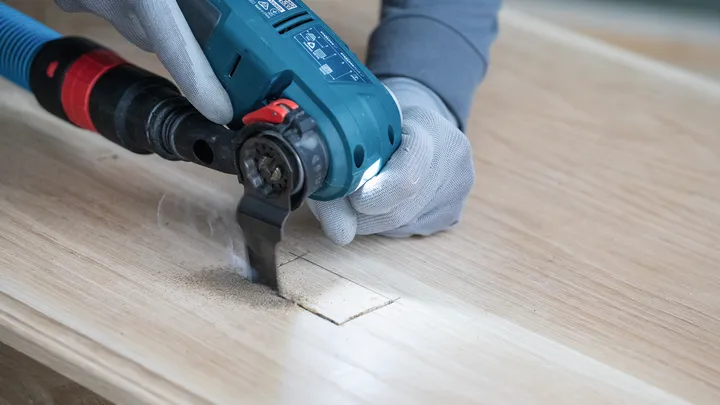 Person wearing safety equipment cuts a rectangular hole in wood with a power tool.