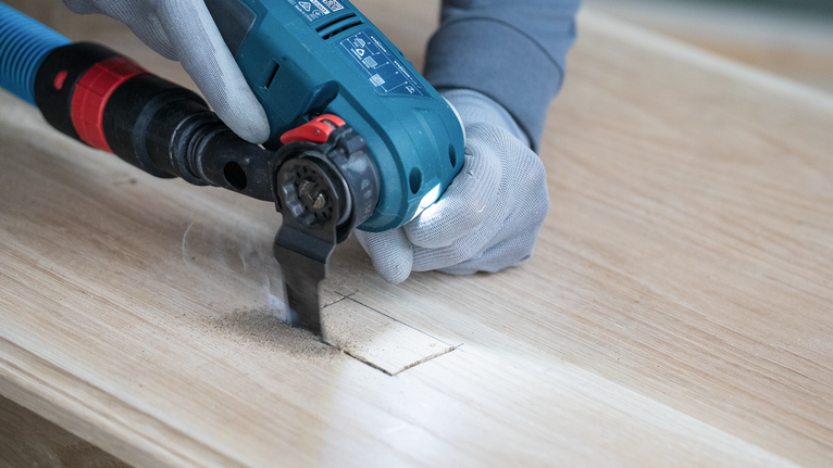 Person wearing safety equipment cuts a rectangular hole in wood with a power tool.