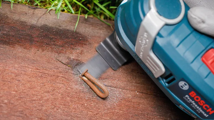 A person uses a power tool to cut through a wooden surface near a metal staple.