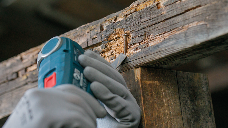 Person wearing safety equipment uses a power tool to remove decayed wood from a beam.