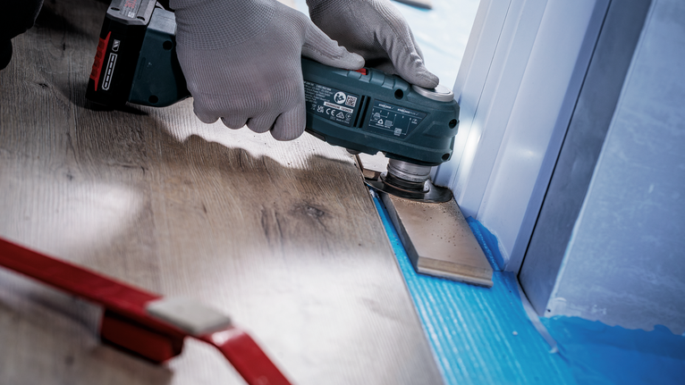 Person wearing safety equipment trims flooring near a door frame using a power tool.