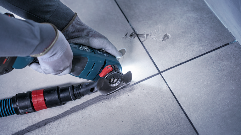 Person wearing safety equipment uses a power tool to cut along a tile seam.