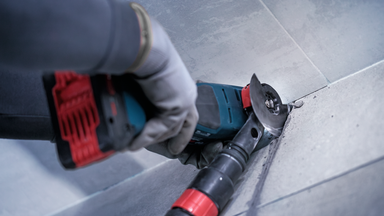 A person wearing safety equipment cuts tile grout in a corner with a power tool.