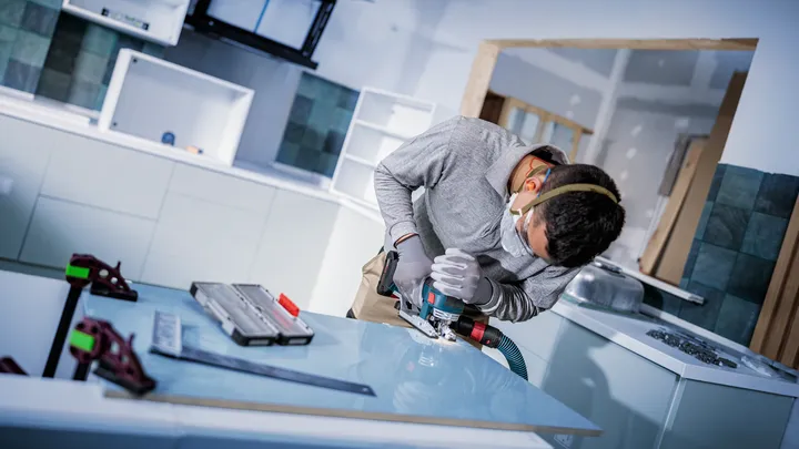 Person wearing safety equipment drills into a glass panel in a kitchen workspace.