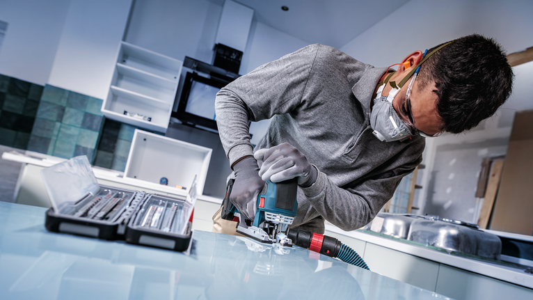 Person wearing safety equipment uses a power jigsaw to cut a panel in a kitchen.