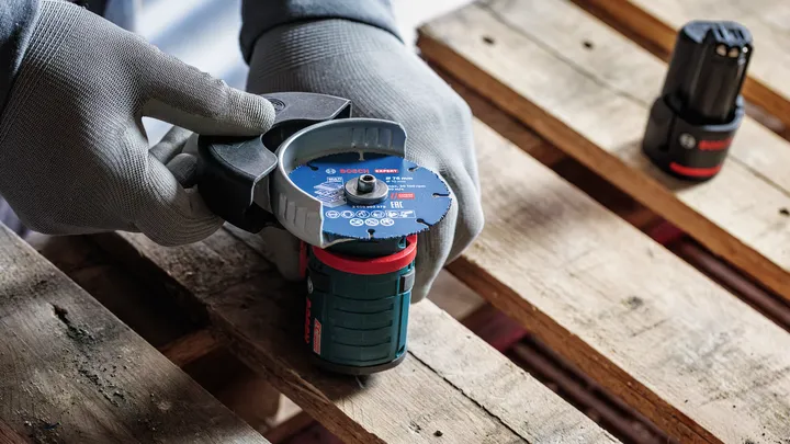 Person wearing safety equipment holds a cordless angle grinder on a wooden pallet.