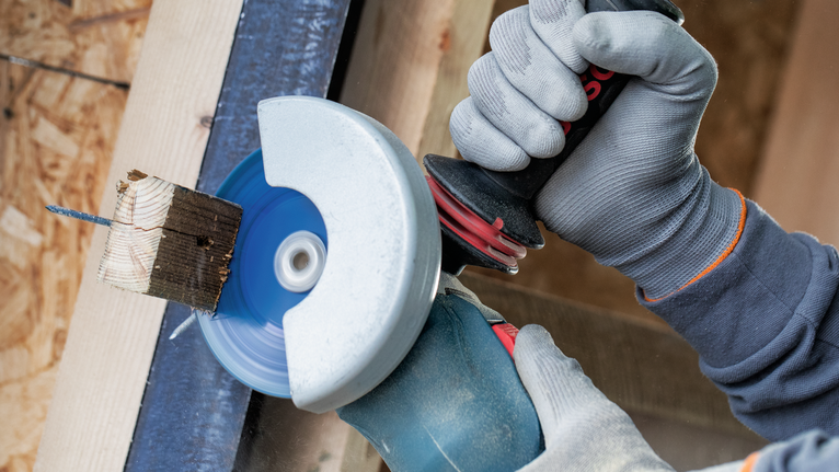 Person wearing safety equipment uses an angle grinder to cut a wooden block with a nail.